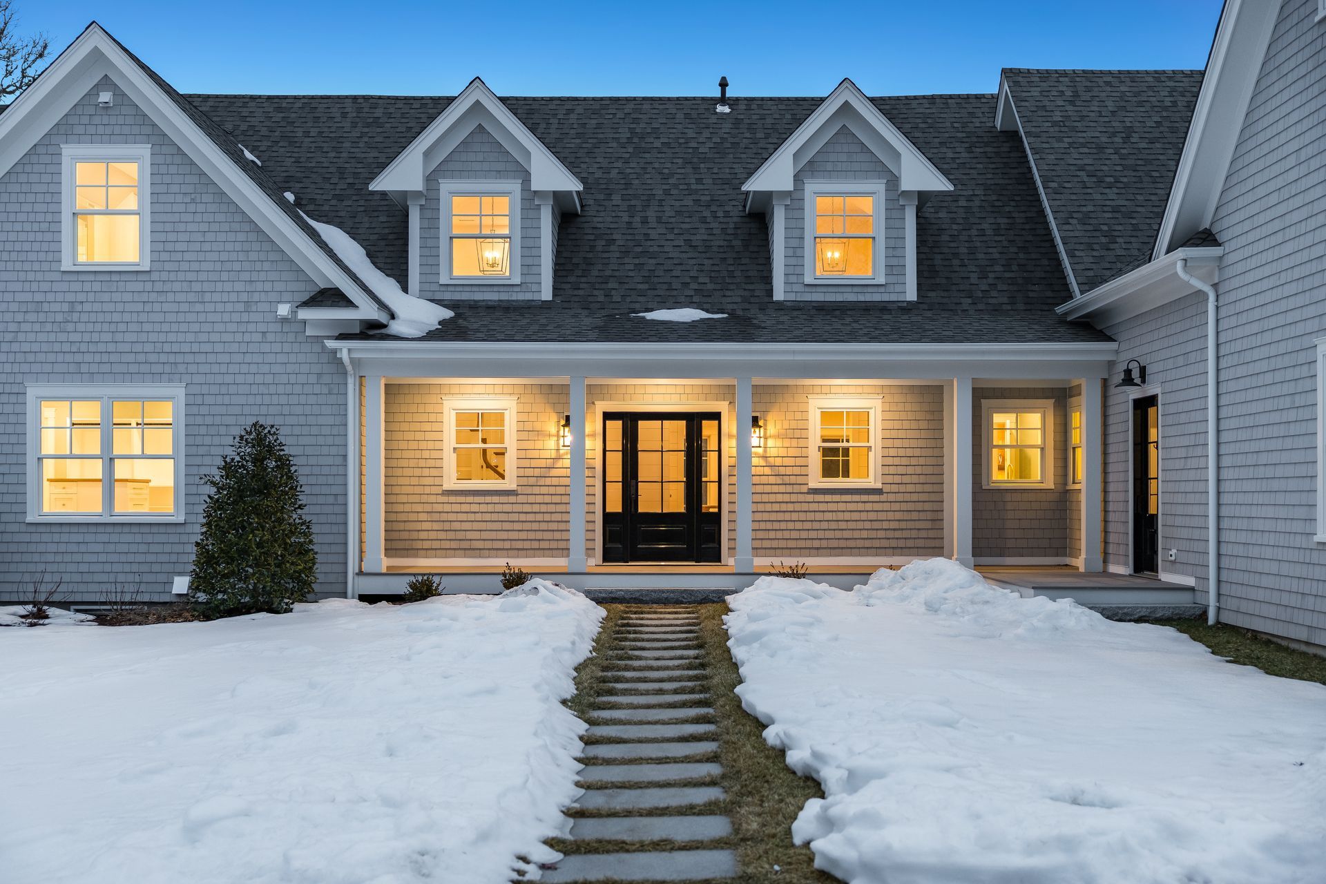 A grey, two-story house with white trim, a covered porch, and dormer windows, set in a snowy landscape at twilight.