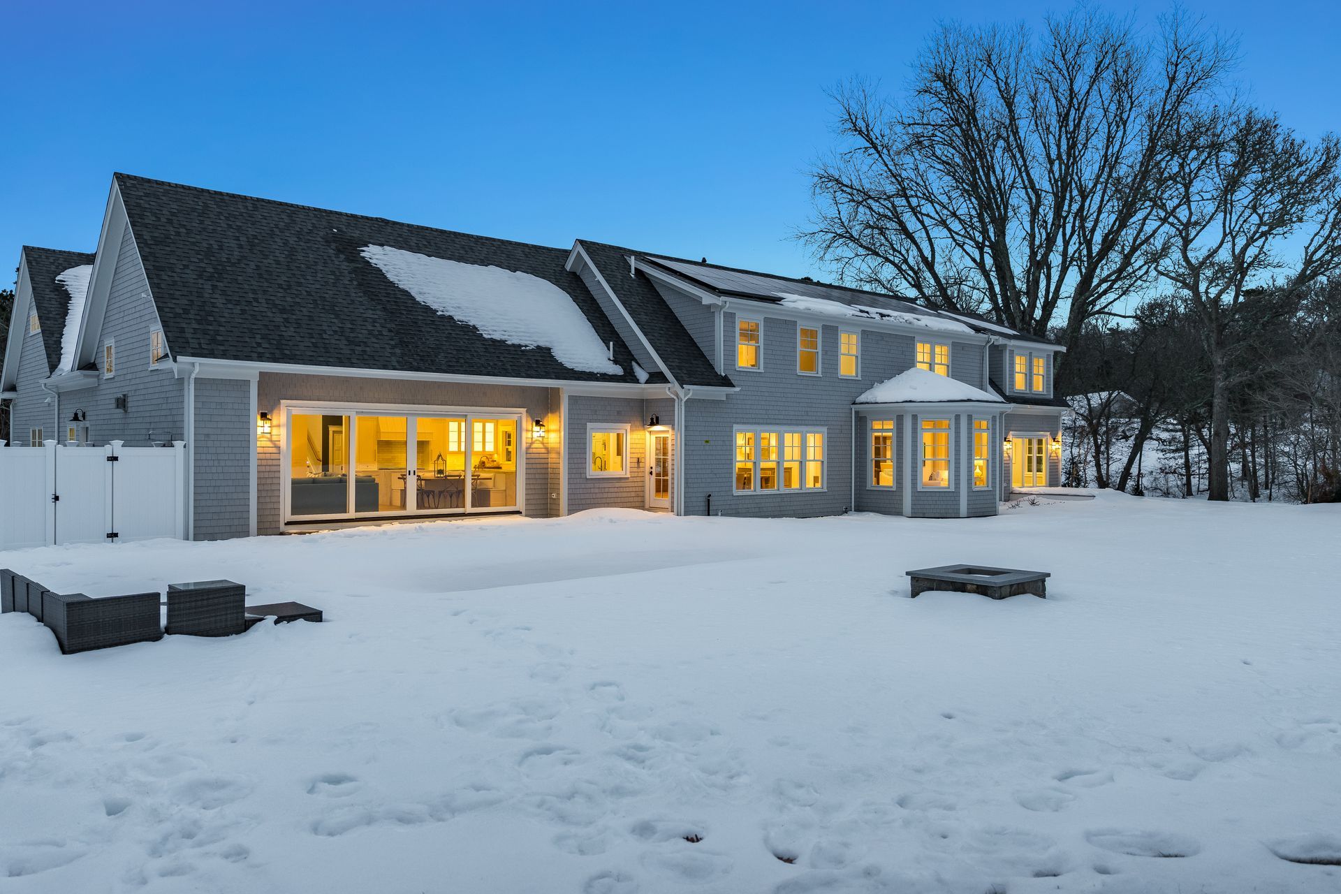 A two-story grey house at dusk, illuminated by interior lights, with snow covering the roof and a large backyard.