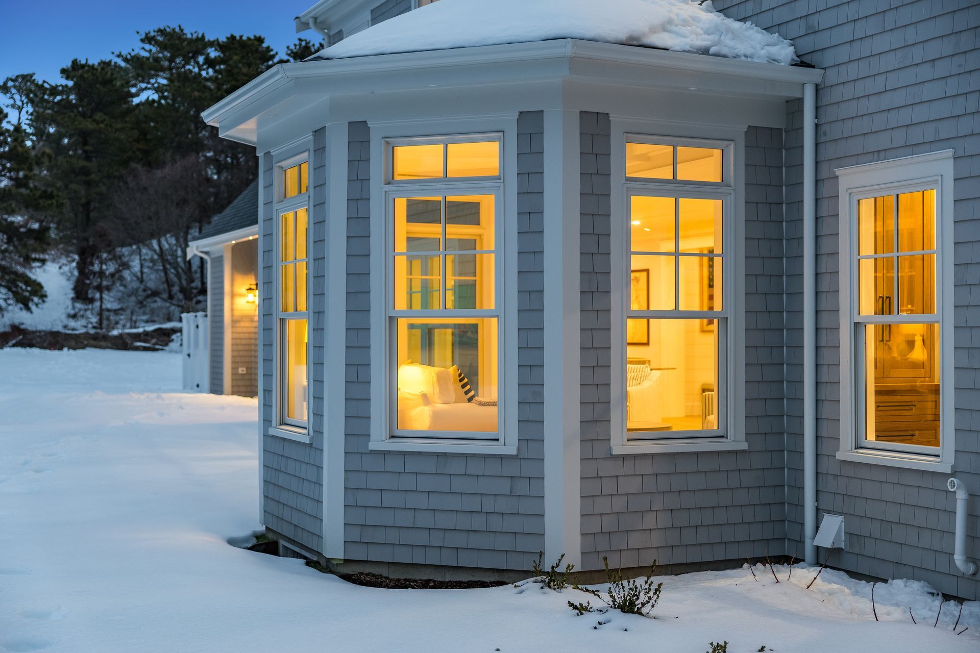 Exterior view of a gray-shingled home at twilight with warm, glowing windows overlooking a snow-covered yard.