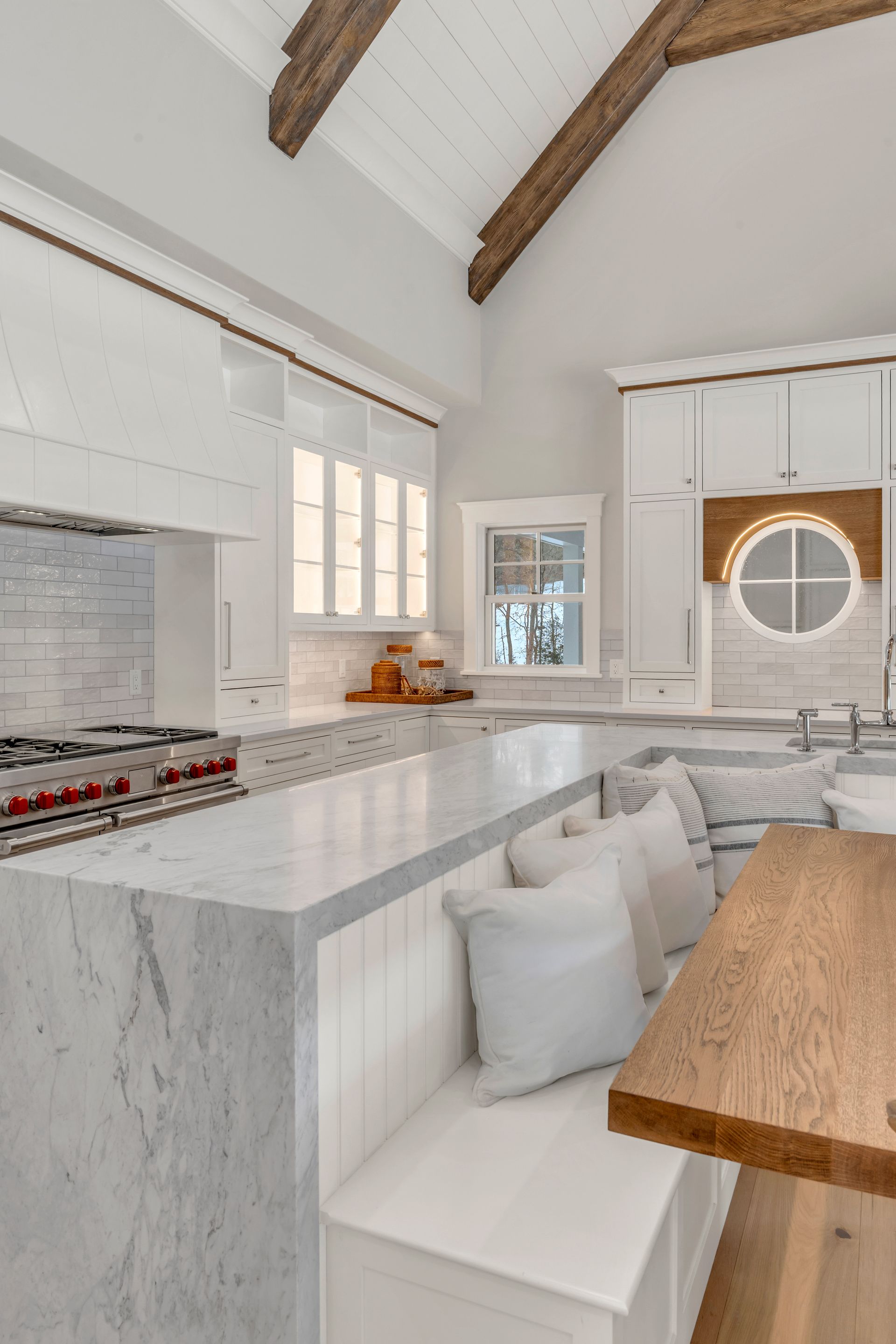 A modern white kitchen featuring a marble island with built-in booth seating, a wooden table, and exposed wood beams.