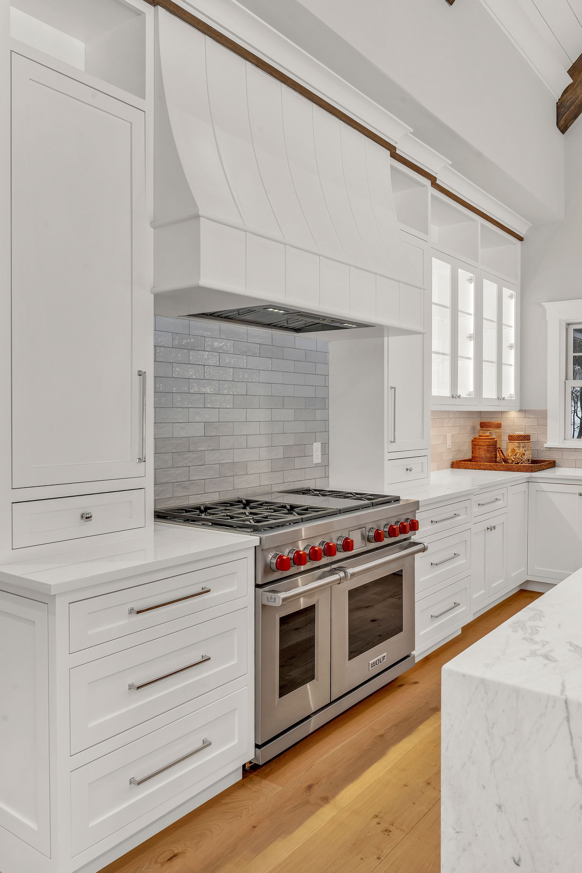 A modern white kitchen featuring a gas range, a custom range hood, gray tile backsplash, and light wood flooring.
