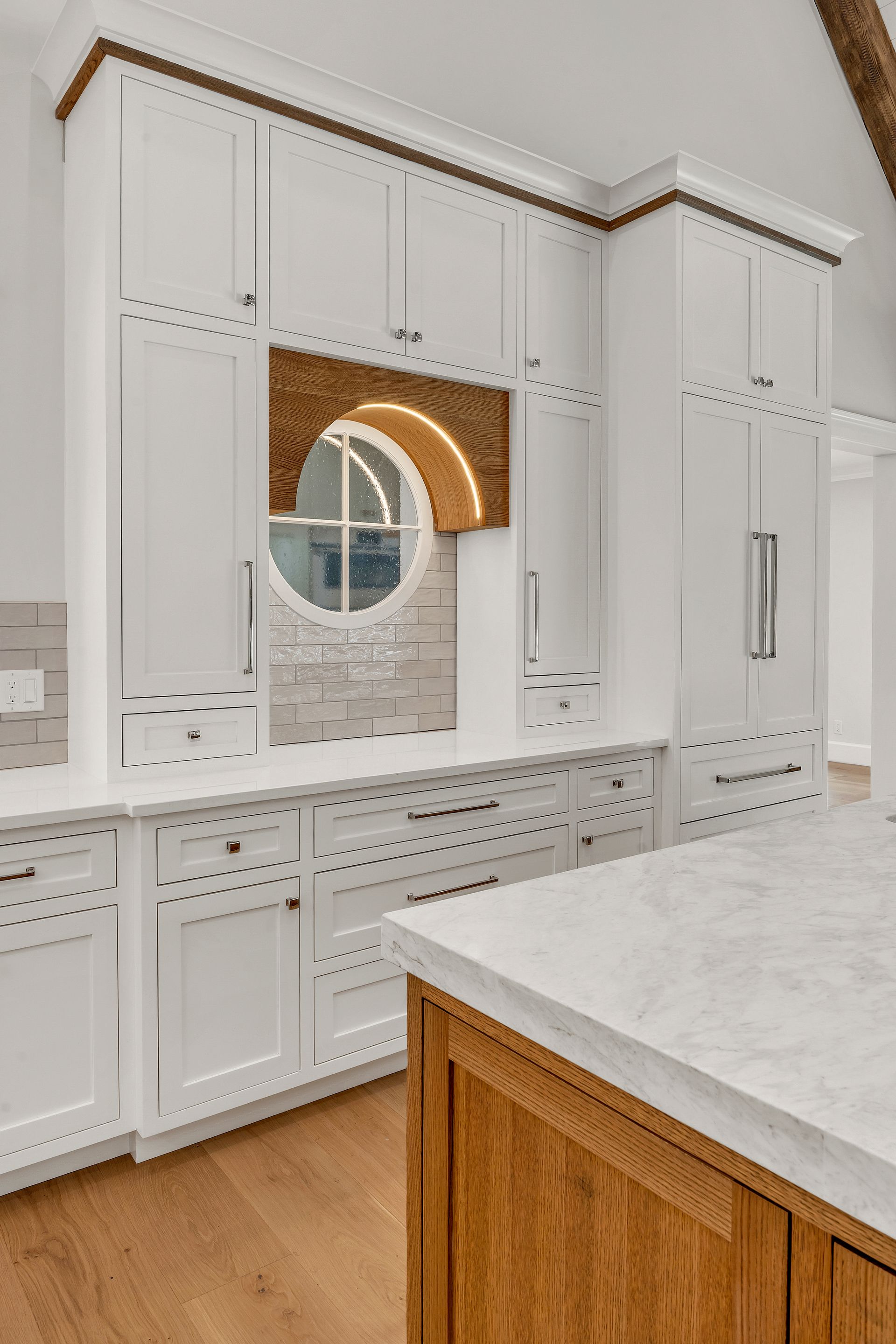 A modern kitchen with white cabinetry, a wood-accented arched window nook, and a light-toned marble island countertop.