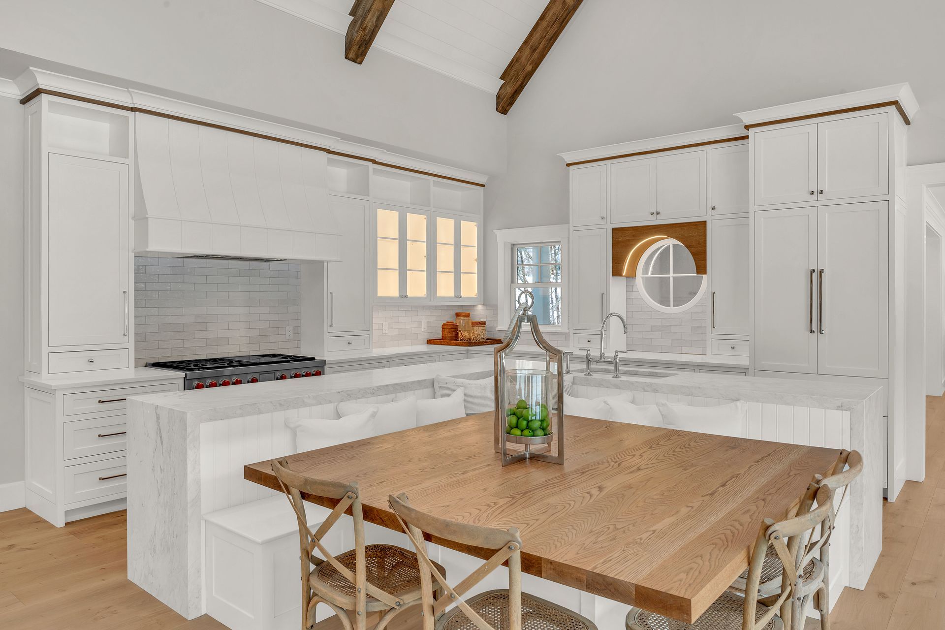 A bright, modern white kitchen featuring a large wooden dining island, a gas stove, and exposed wooden ceiling beams.