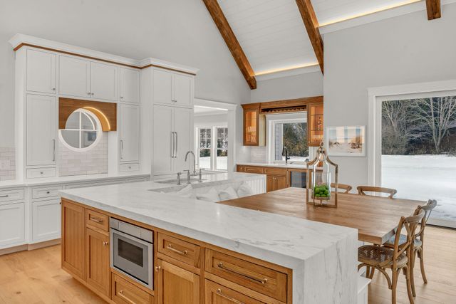 Bright kitchen with a large marble island, wood cabinets, vaulted ceiling with exposed beams, and a view of snow outside.
