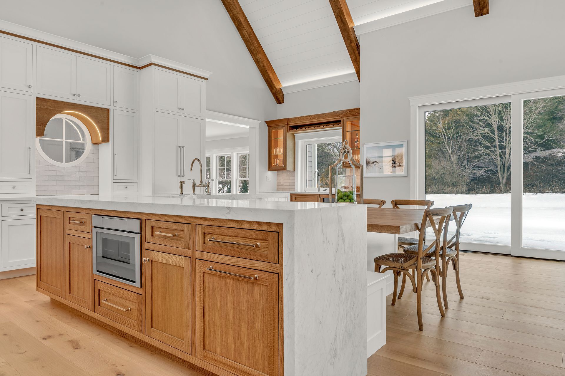 A white and wood kitchen featuring a large marble-topped island with built-in oven, vaulted ceilings, and dining area.