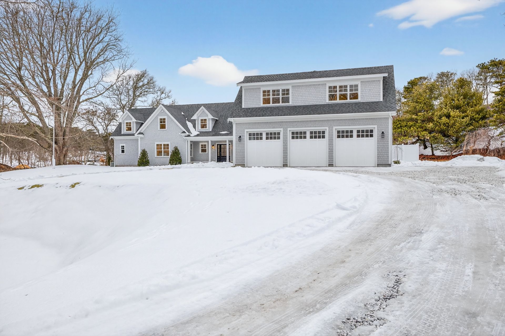 A gray, two-story house with a three-car garage and dormer windows, surrounded by a snow-covered yard and a gravel drive.