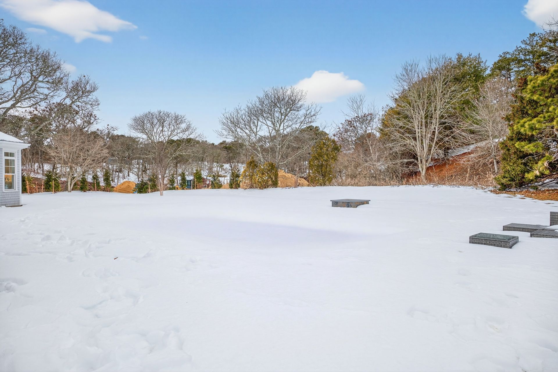 A snow-covered backyard on a sunny day, featuring a corner of a building, scattered patio tiles, and bare trees in the back.