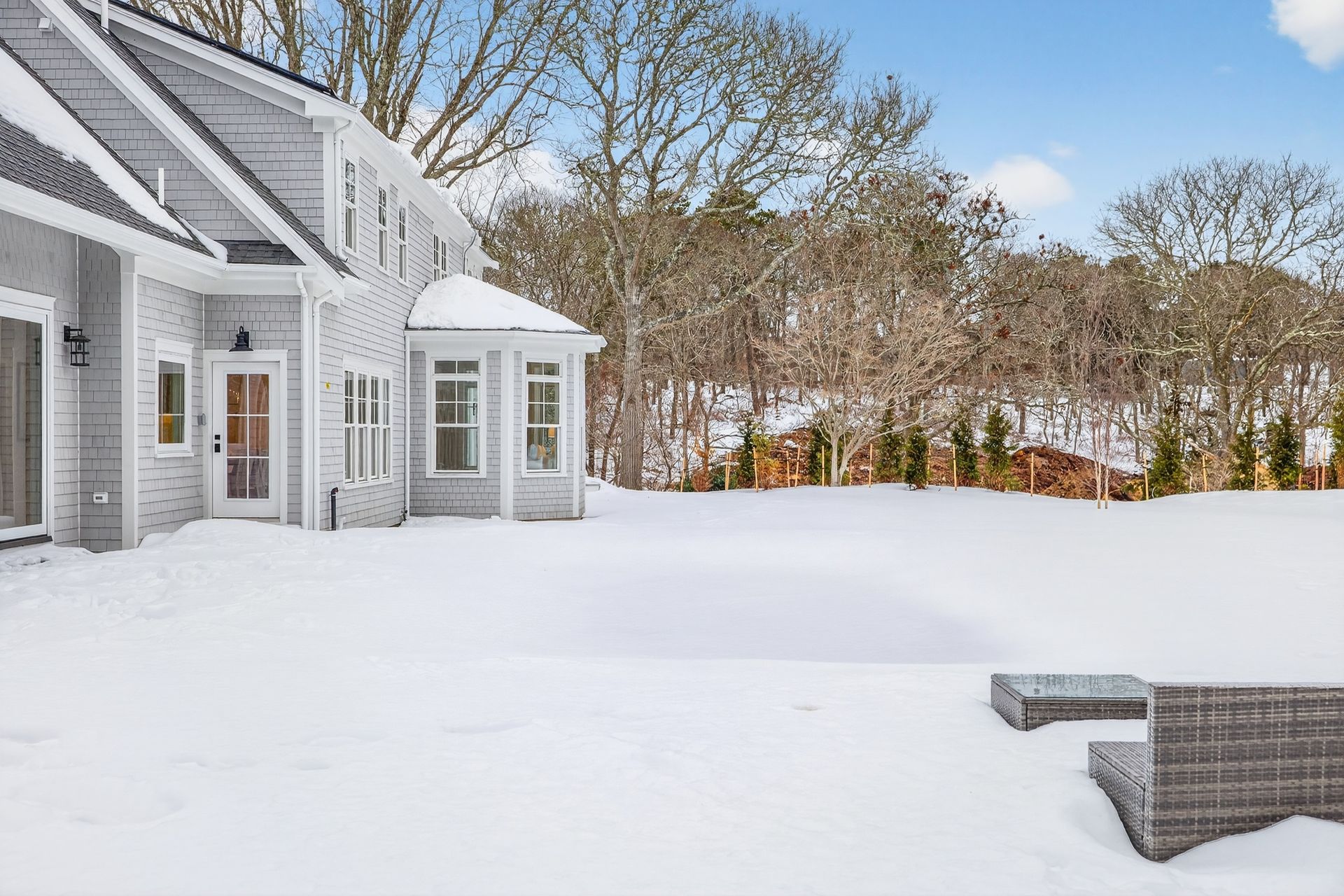 A gray house sits in a snow-covered yard with bare trees in the background and outdoor furniture on a patio.