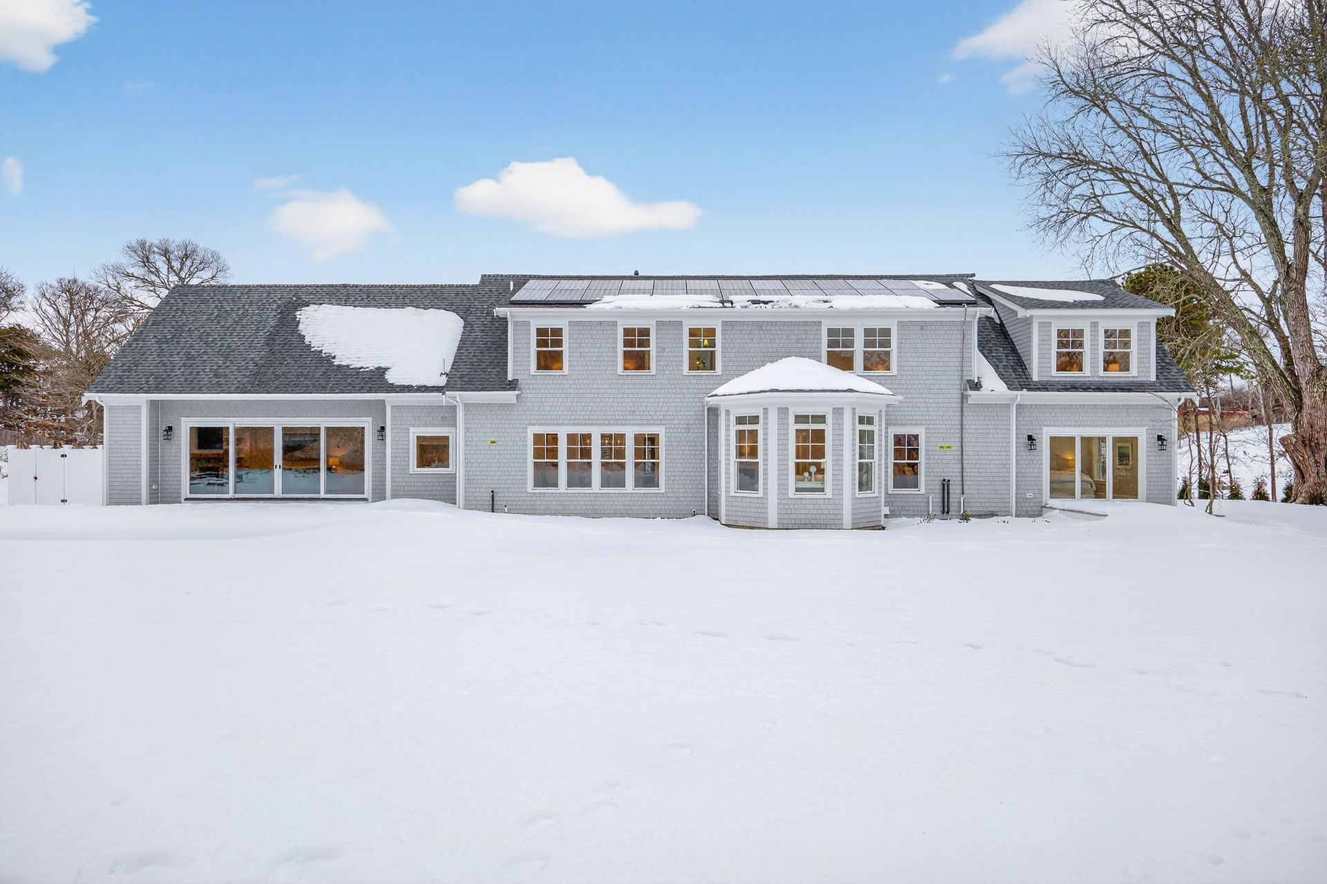 The rear exterior of a gray, two-story house with a snow-covered yard and roof under a clear blue sky.