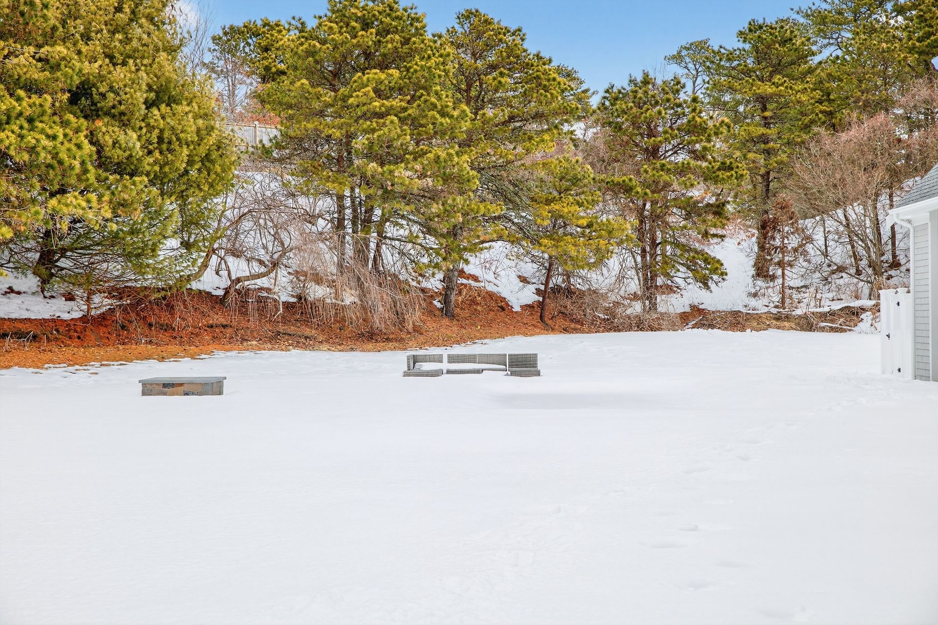 A snow-covered backyard with a rectangular fire pit and a modern wooden sectional bench near a line of trees.