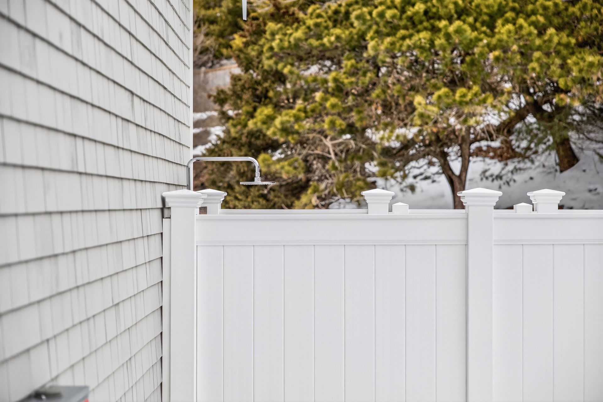 A white vinyl privacy fence stands next to a gray shingled building with a shower fixture visible above the fence.