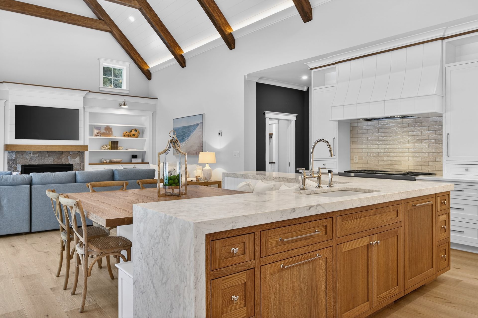 A bright, open-concept kitchen with a large marble waterfall island, natural wood cabinets, and vaulted beamed ceilings.
