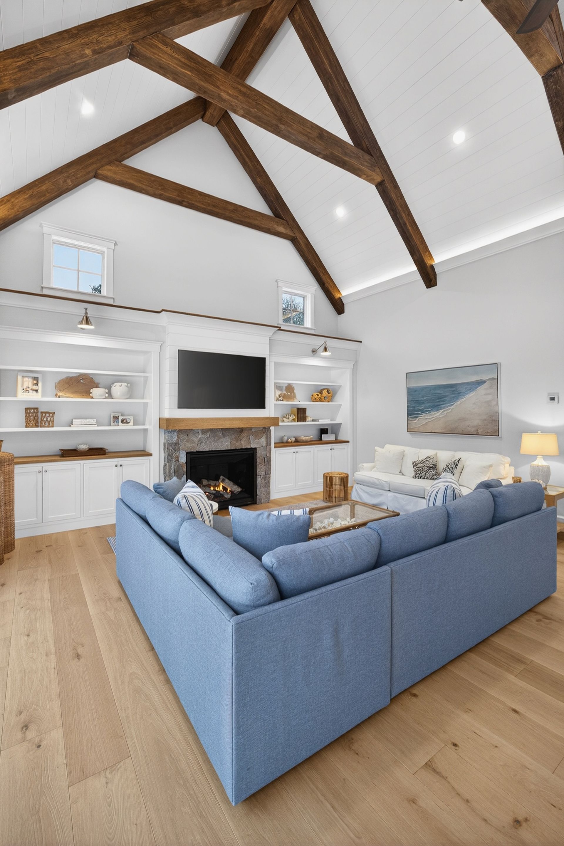 Living room with wood beams, white walls, stone fireplace, blue sectional sofa, and hardwood floors.