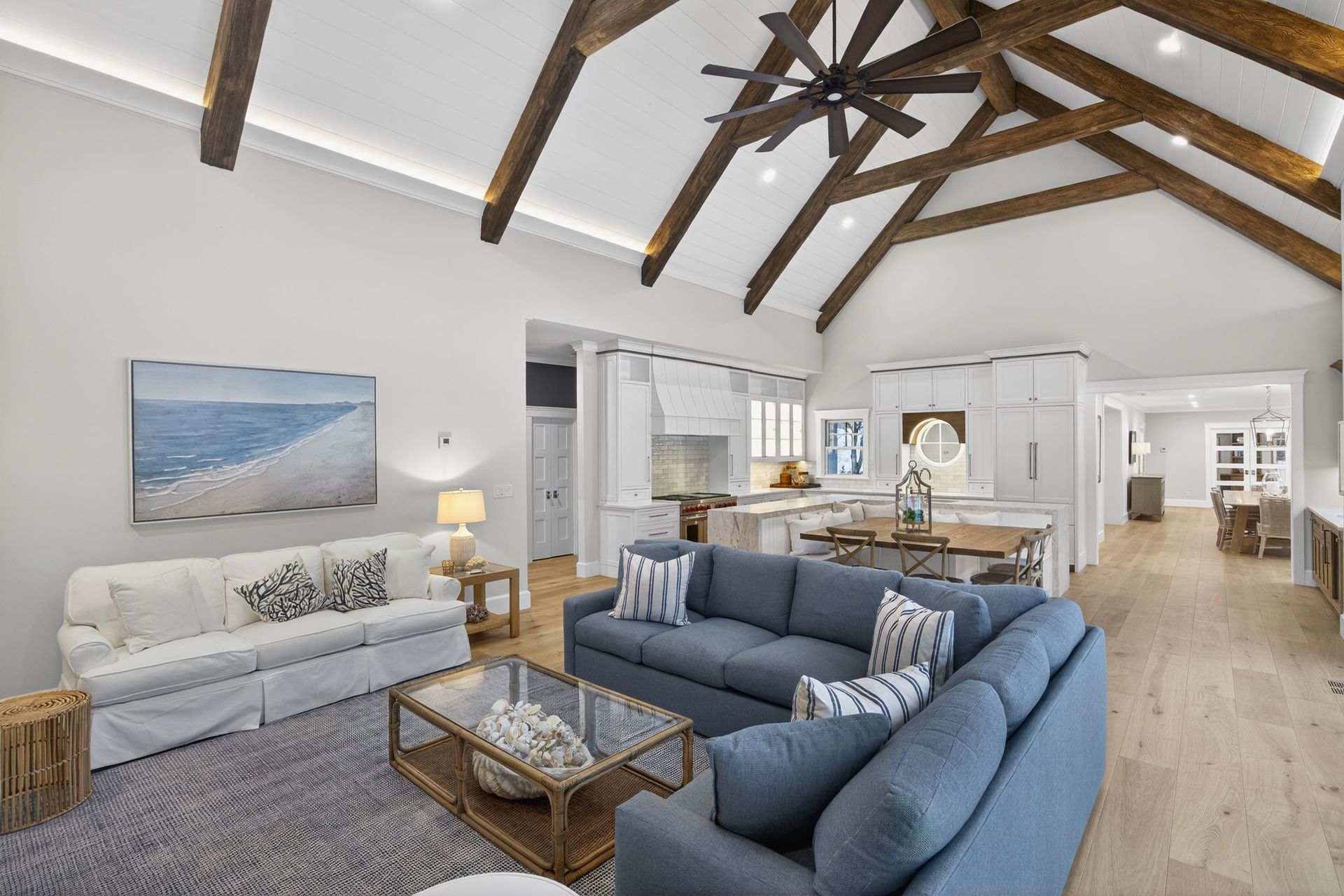 Living room with vaulted ceiling, exposed beams, blue and white furniture, hardwood floors, and open kitchen.
