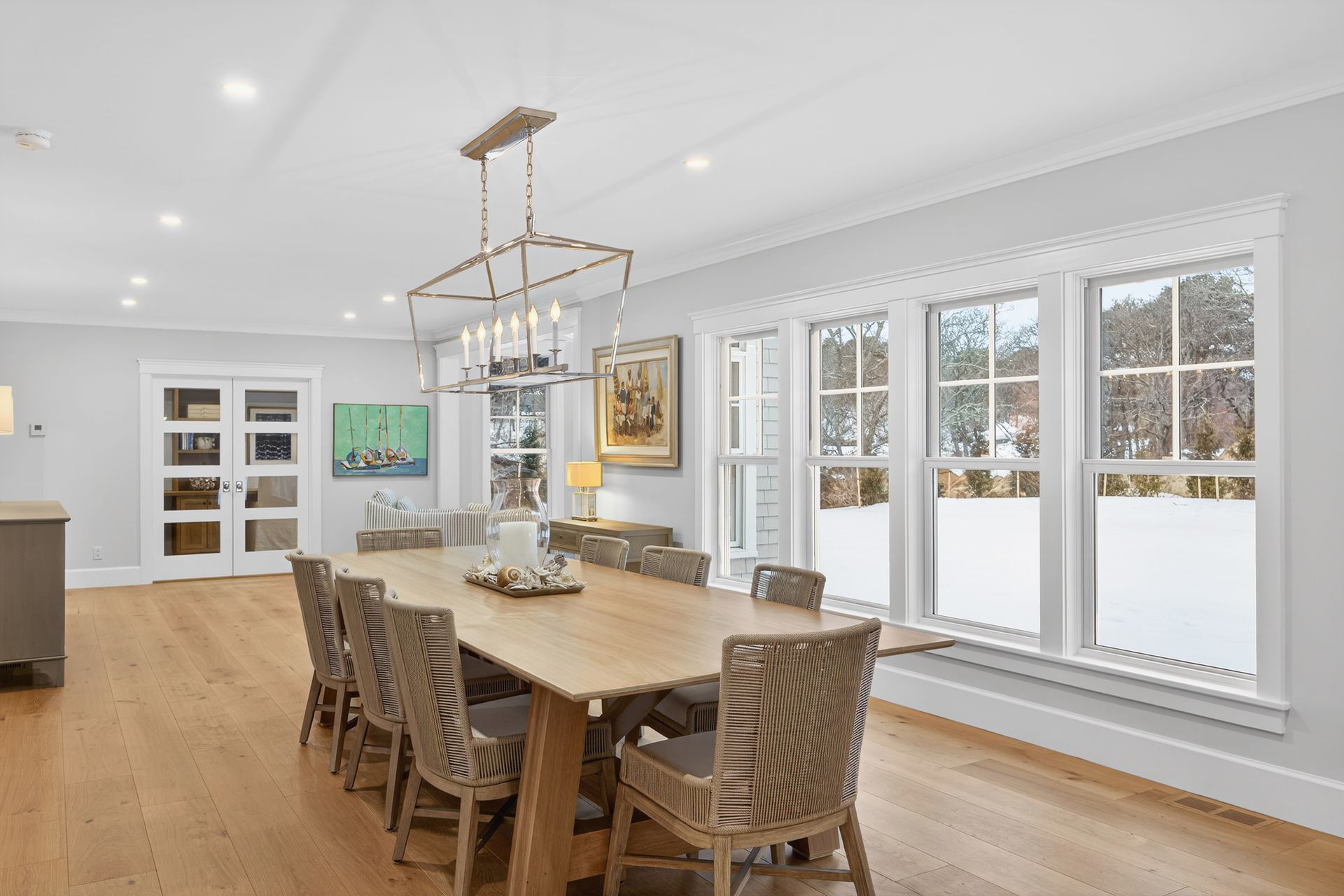 Dining room with long wooden table, wicker chairs, windows, and chandelier.