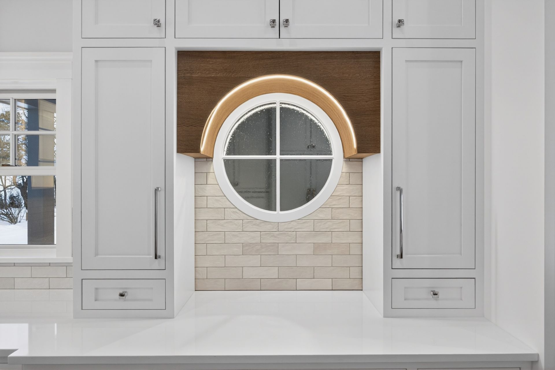 Kitchen with white cabinets flanking a round window with a brick wall backdrop.