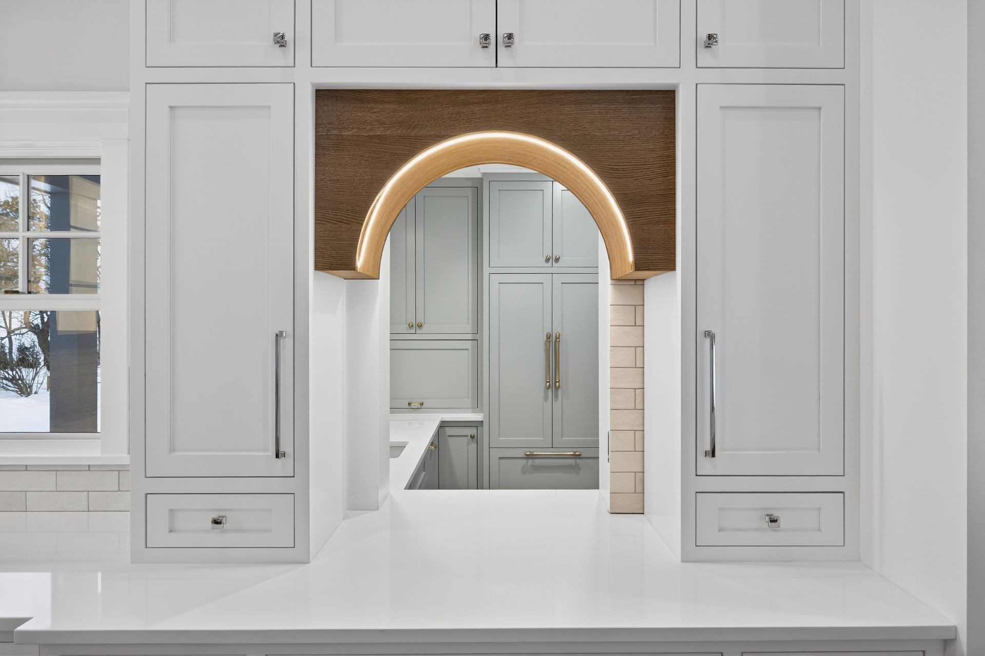 White kitchen with archway revealing another room with cabinets. Brown arch with glowing lights.