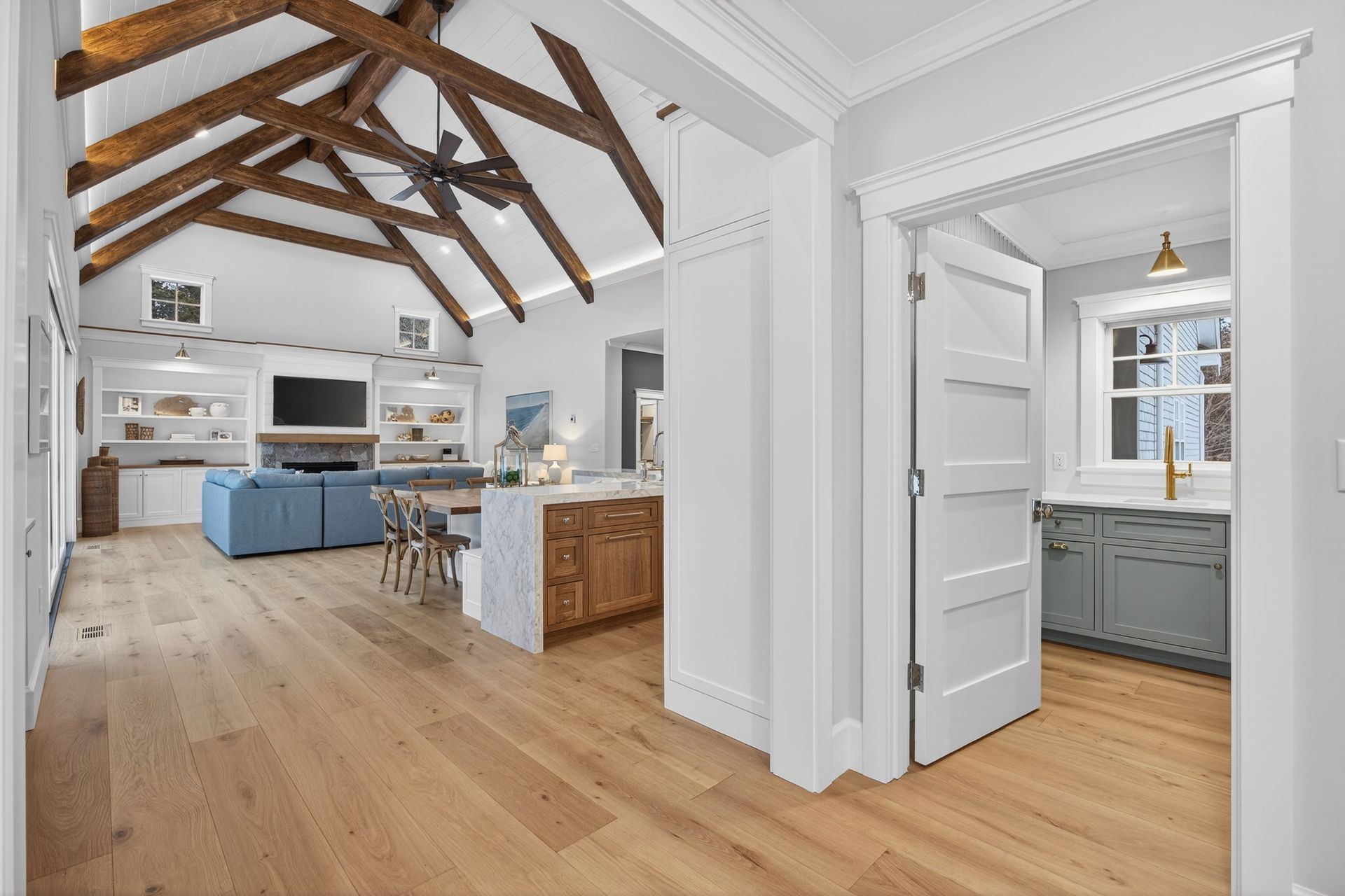 Open-concept living space with light wood floors, a kitchen island, and vaulted ceiling with exposed wooden beams.