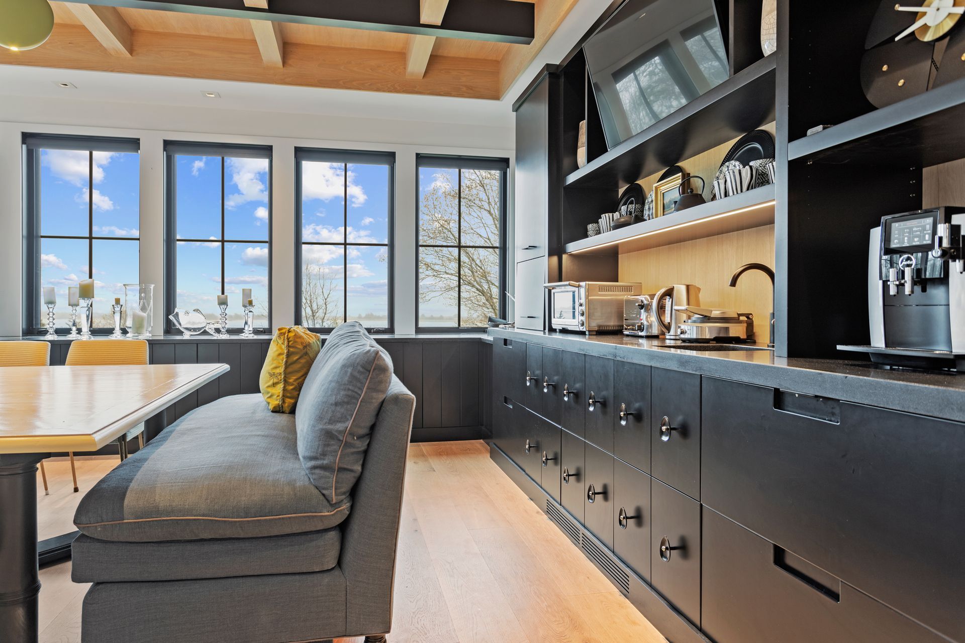 A modern kitchen featuring black cabinetry, a built-in bench with a gray sofa, and a table beneath large windows.