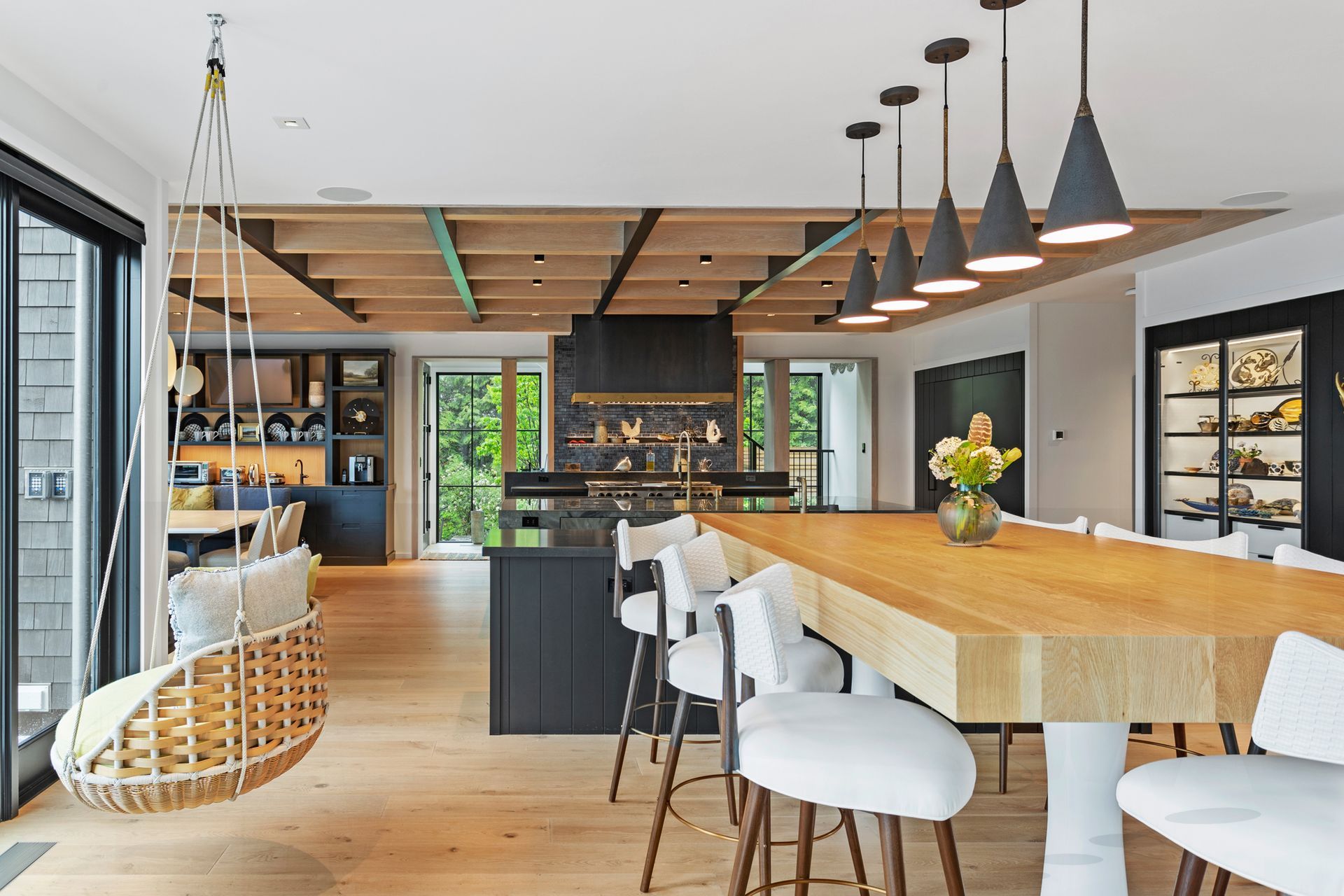 A modern kitchen and dining area featuring a large wooden island with white chairs, hanging lights, and a rattan swing chair.
