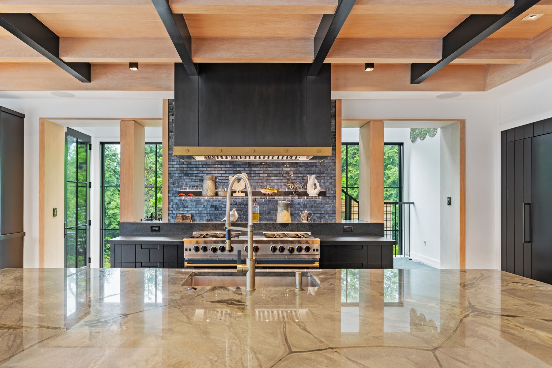 A modern kitchen with wood-beamed ceilings, a large marble island, dark cabinets, and a blue tile backsplash.