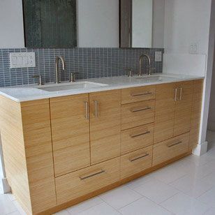 A double-sink bathroom vanity with light wood cabinets, silver hardware, and a grey mosaic tile backsplash.