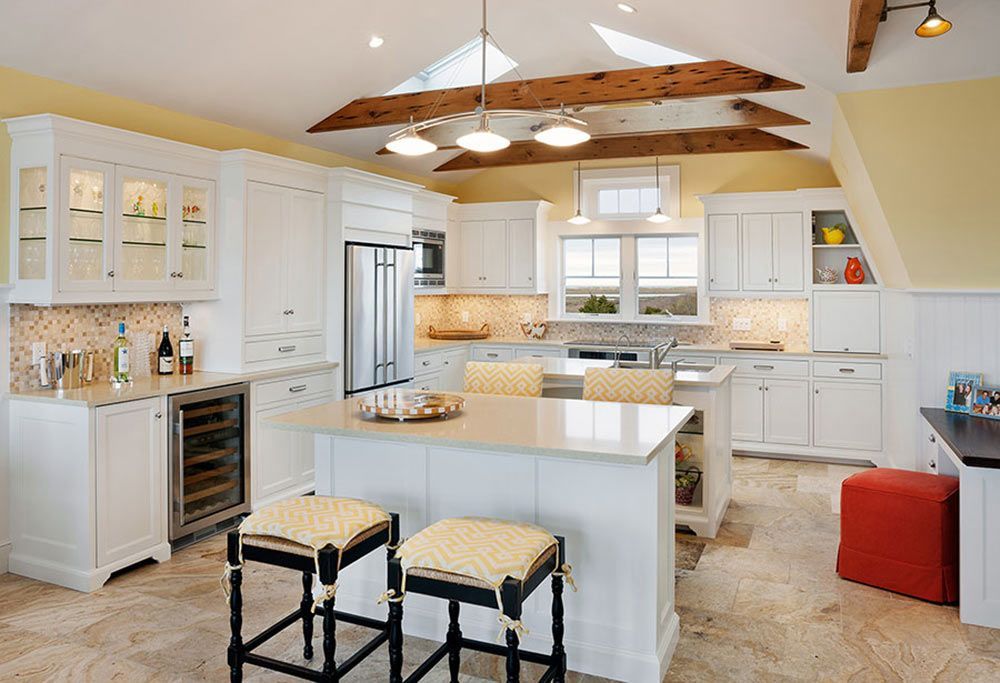 White kitchen with an island, two stools, light-colored stone floors, and wooden beams on a vaulted yellow ceiling.