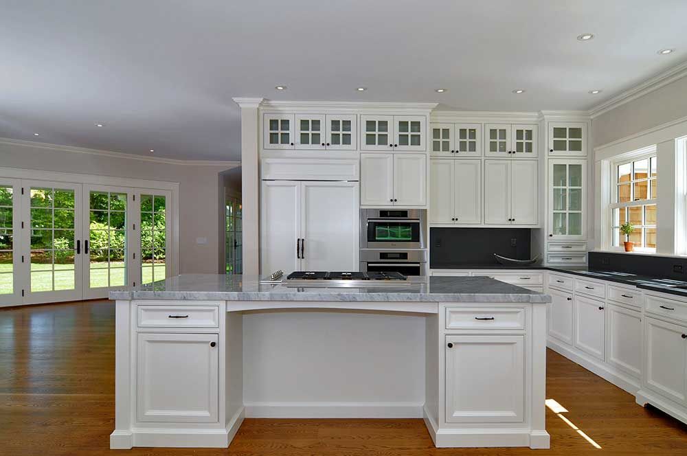 A modern kitchen featuring white cabinets, a large marble-topped island, a built-in oven, and a view of a garden.