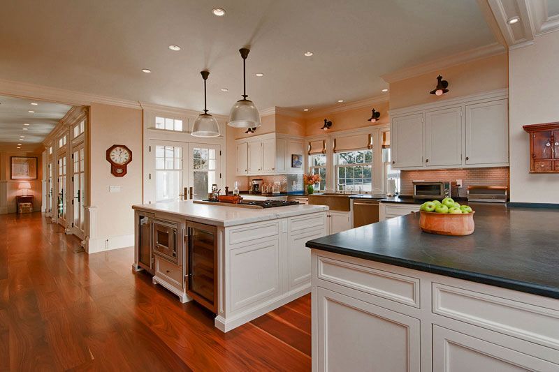 A bright kitchen with white cabinets, a large central island, dark countertops, and polished wood floors.