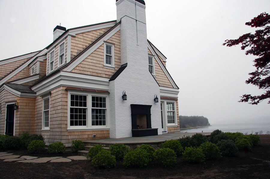 A shingled house with a white brick chimney and outdoor fireplace sits on a coast by a foggy ocean.