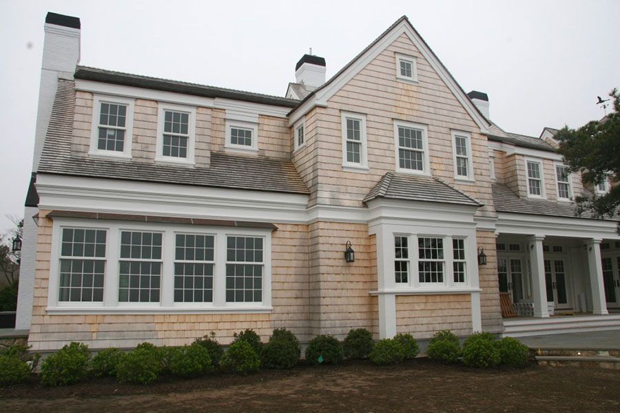 A light-colored house with shingle siding, white trim, multiple windows, and a dark shingled roof under a gray sky.