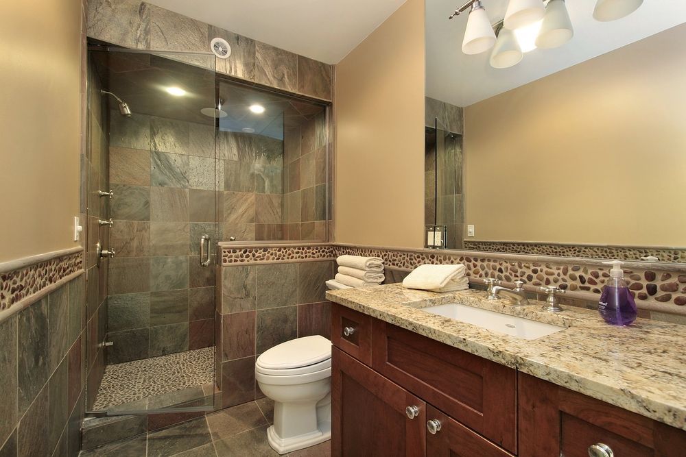 A bathroom with a stone-tiled shower, a white toilet, and a vanity featuring a granite countertop and dark wood cabinets.