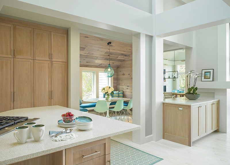 A bright modern kitchen with light wood cabinets, a stone island, and a dining area with blue accents visible beyond.
