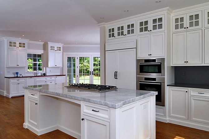 A bright, spacious kitchen featuring white cabinetry, a marble-topped island with a cooktop, and stainless steel appliances.
