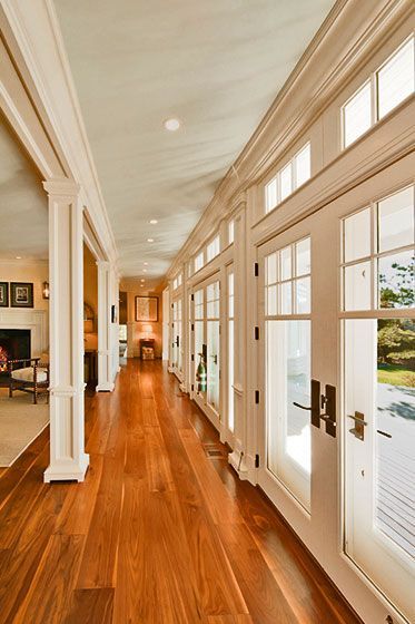 A sunlit hallway with polished wood floors, white columns, and a row of glass doors looking out to a garden.