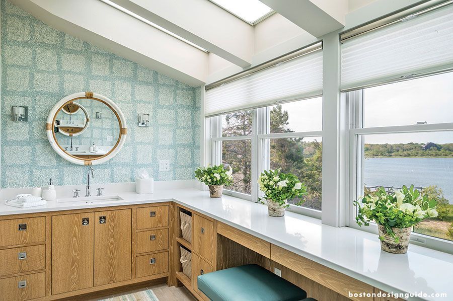 A bright, light-filled bathroom with oak vanity cabinets, a round mirror, patterned blue wallpaper, and a water view.
