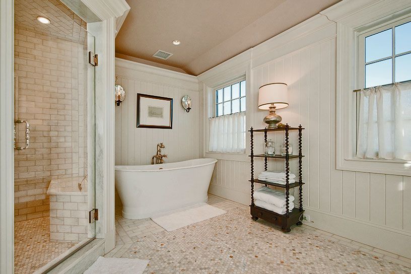 A bright, elegant bathroom with a freestanding white tub, tiled shower, pedestal lamp on a shelf, and marble-flecked floors.