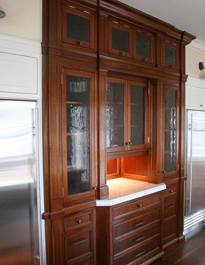 A tall, dark wood kitchen hutch with glass-front cabinets, a white countertop, and drawers between two stainless fridges.