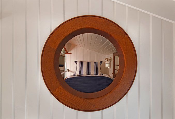 A round, wooden-framed mirror mounted on a white paneled wall reflects a bedroom with striped pillows and a blue blanket.
