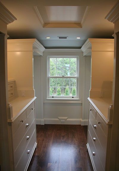 A walk-in closet with white cabinets, dark hardwood floors, and a window with a view of green trees.