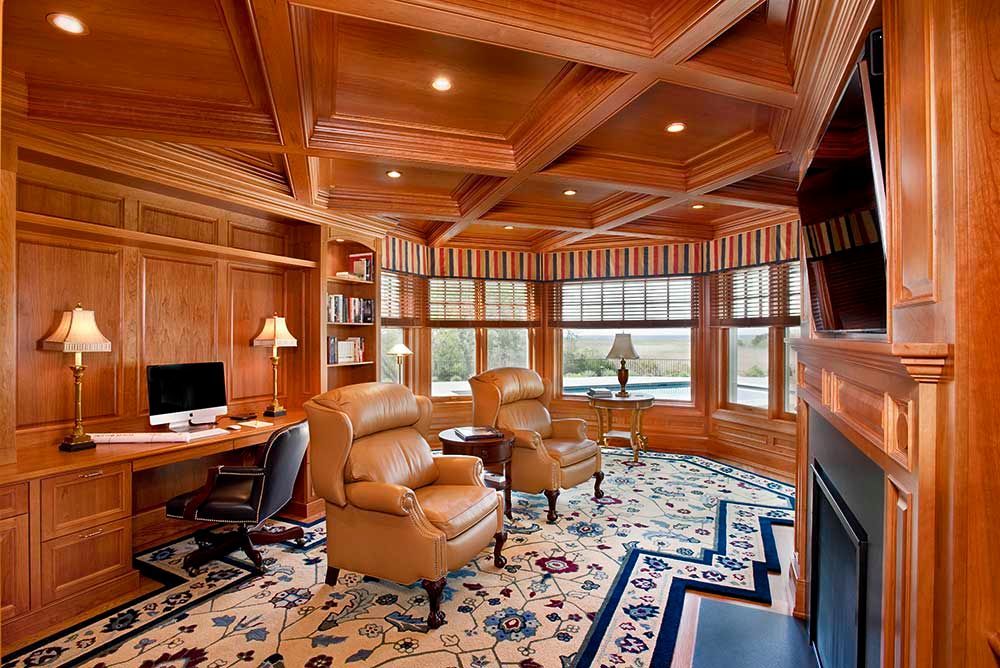 A luxurious home office with rich wood paneling, a coffered ceiling, a desk, two leather armchairs, and patterned carpet.