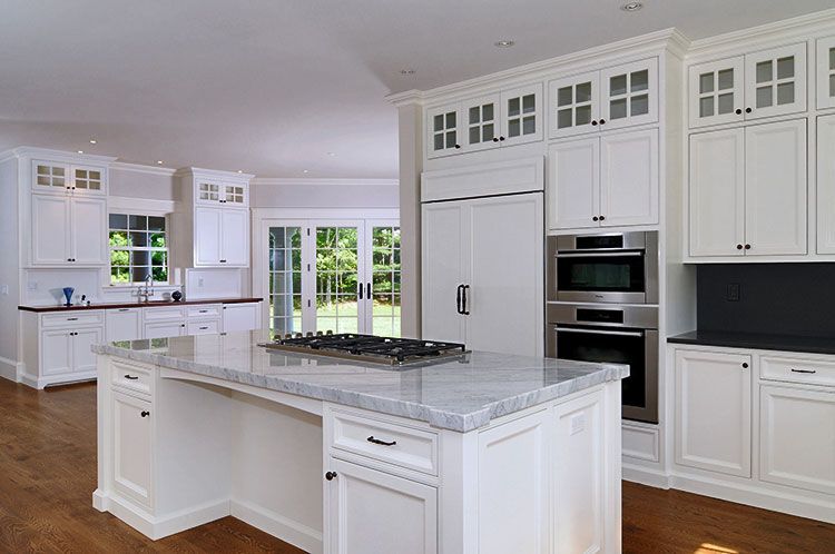 A bright kitchen featuring white cabinets, a large marble island with a stovetop, and stainless steel built-in ovens.
