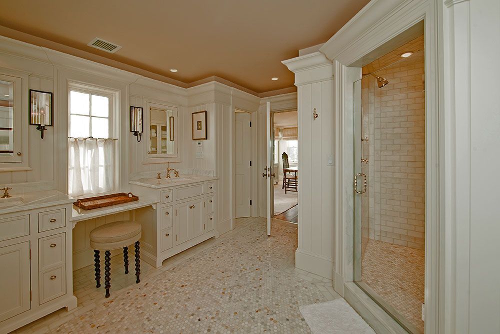 A bright bathroom with white cabinetry, a marble vanity, a stool, and a glass-enclosed shower with tiled walls.