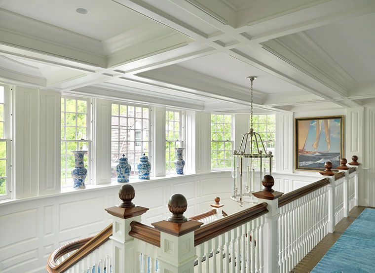 A bright, white-paneled hallway with a wooden staircase railing, coffered ceiling, blue ginger jars, and a sailboat art.