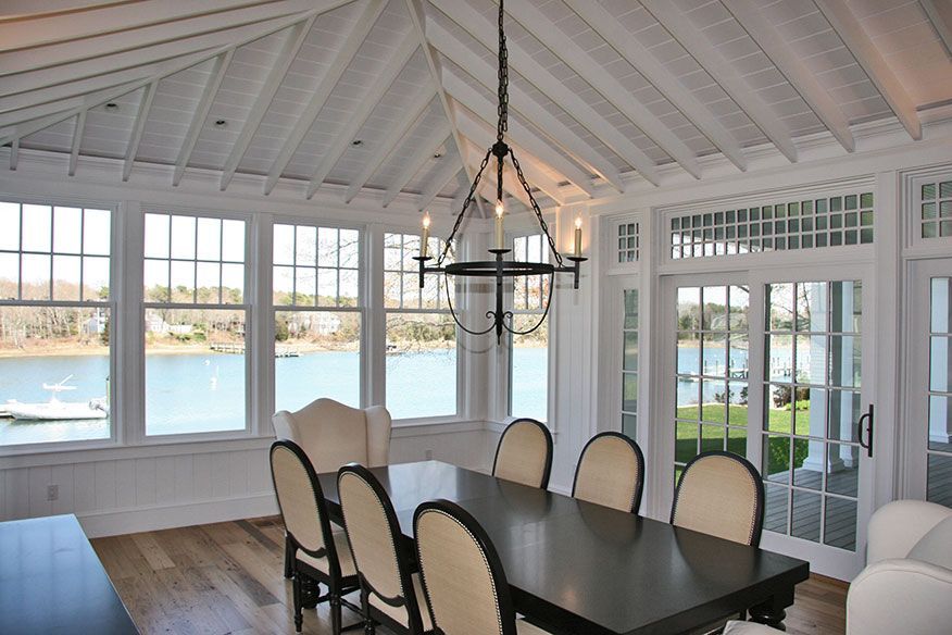 A bright, white dining room with vaulted ceilings, a dark table, and chairs overlooking a calm lake through large windows.