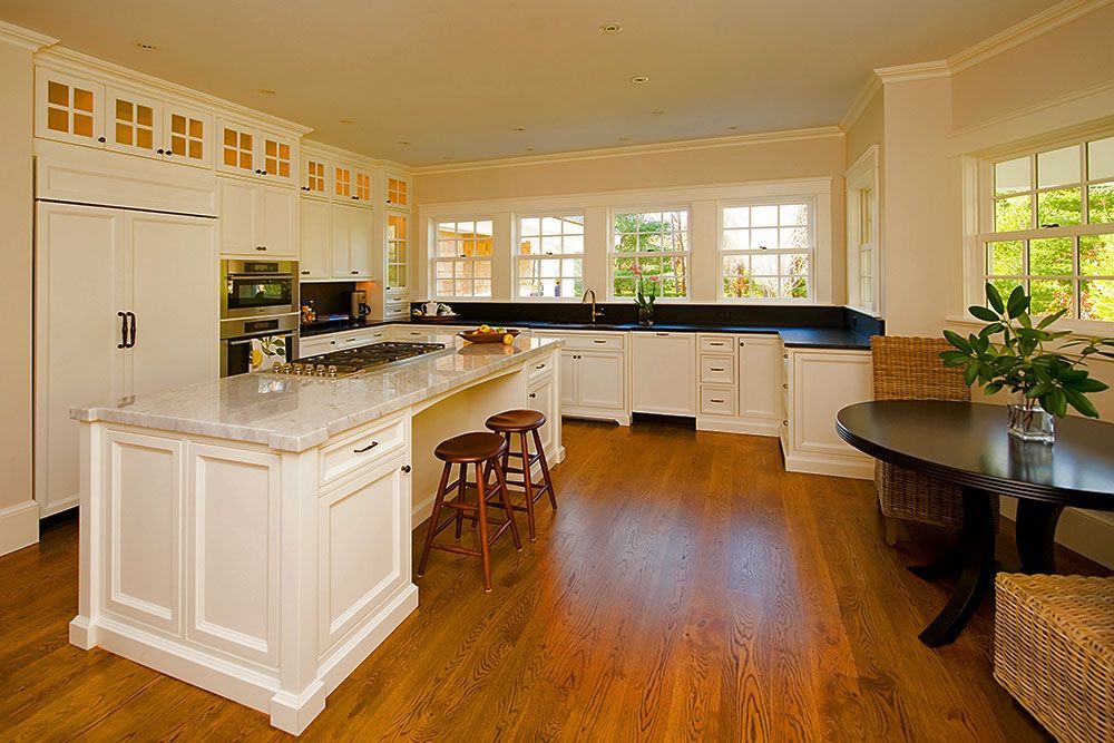 A spacious, bright white kitchen with a marble-topped island, wooden floors, and a dining table by a window.