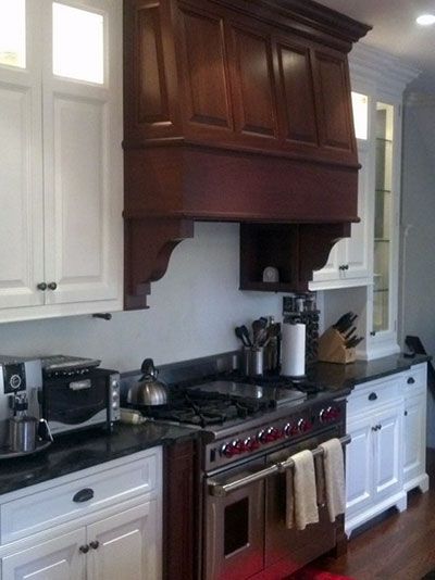 A kitchen features a large, dark wood custom range hood centered above a stainless steel stove between white cabinets.