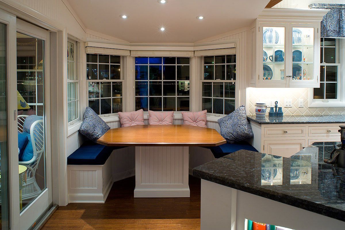 A built-in kitchen banquette with a wooden table, blue cushions, and patterned pillows in a bright, white windowed nook.