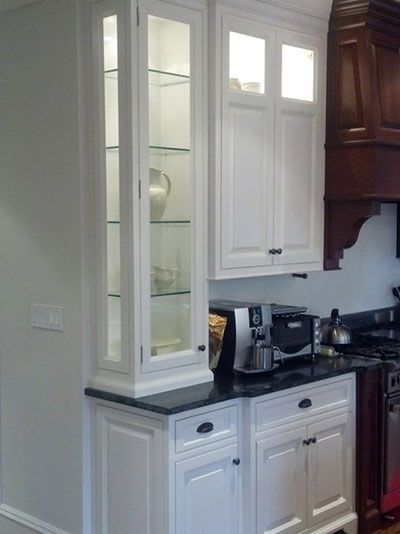 A white kitchen hutch cabinet with glass display doors, glass shelving, and base cabinets with black countertops.