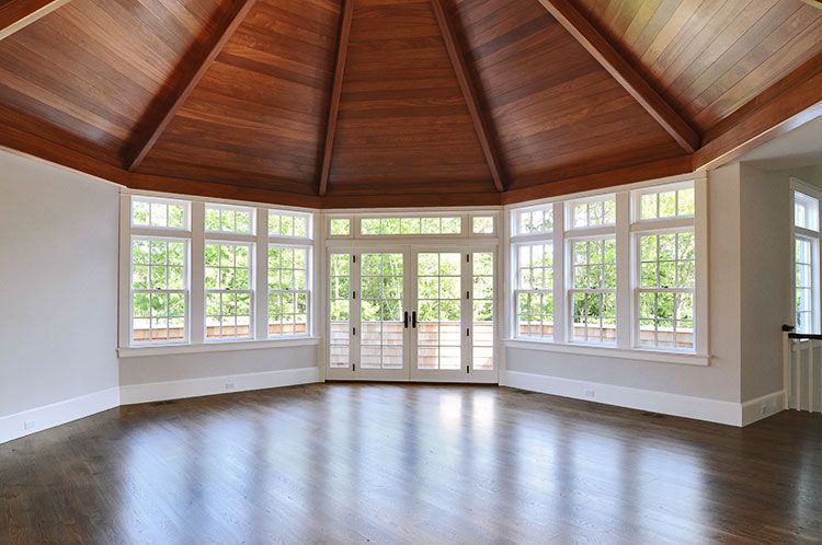 A sunlit, empty room with dark hardwood floors, white walls, large windows, and a vaulted wood-paneled ceiling.